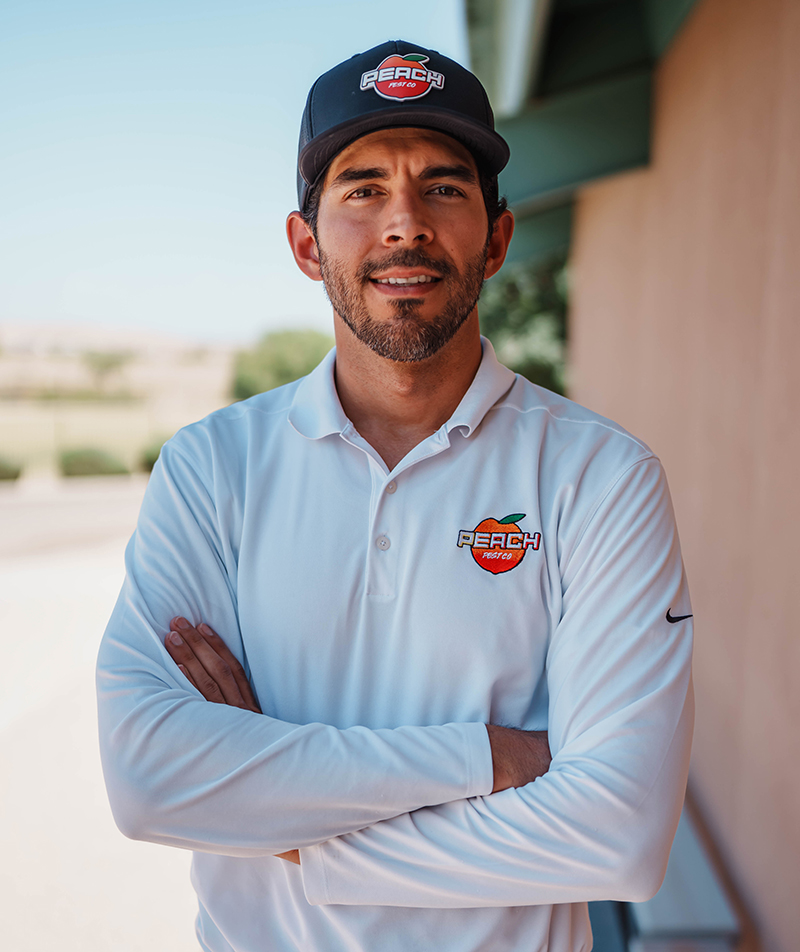 Kadin Hernandez, President and Founder of Peach Pest Co, wearing a branded polo shirt and cap, standing confidently with arms crossed against a sunny outdoor backdrop.